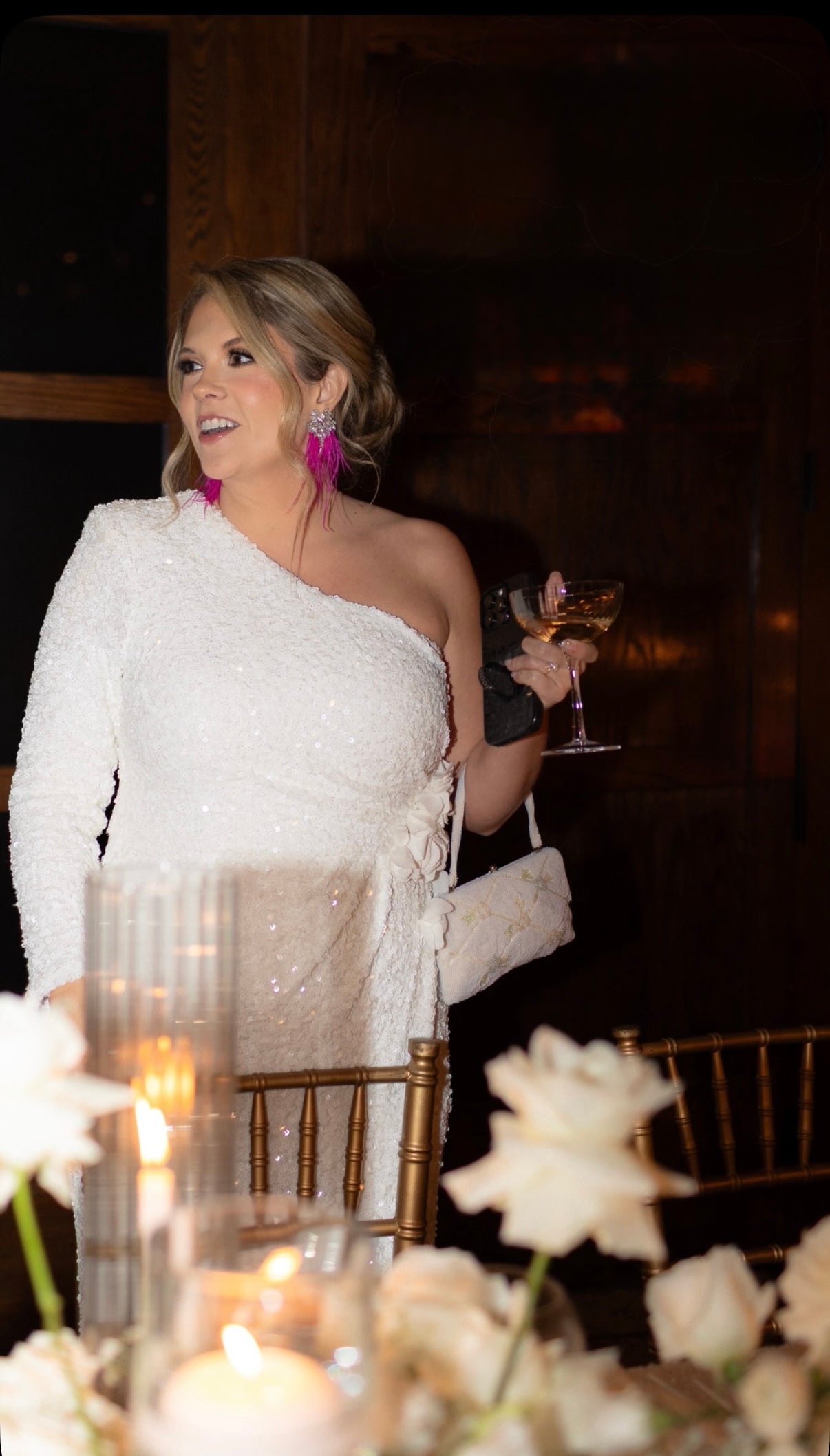 Woman in a white dress and fuchsia feather earrings holding a glass at a formal event with floral decorations.
