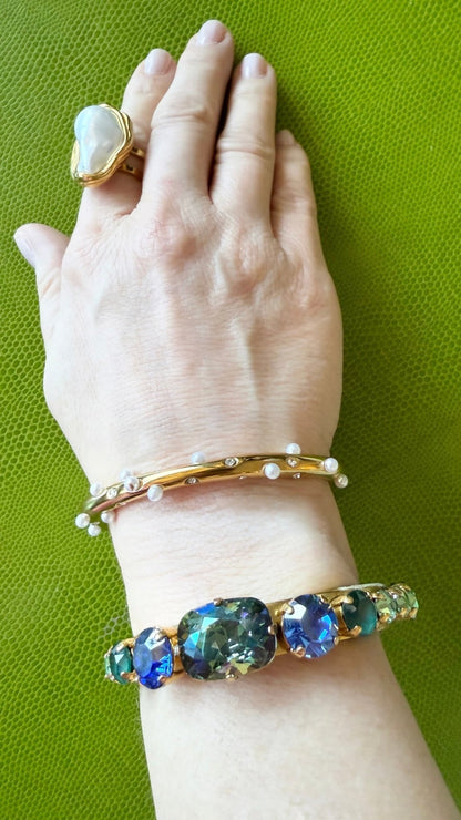 Hand wearing a gold crystal and pearl bracelet, a bracelet with blue gemstones, and a baroque pearl ring on a green background