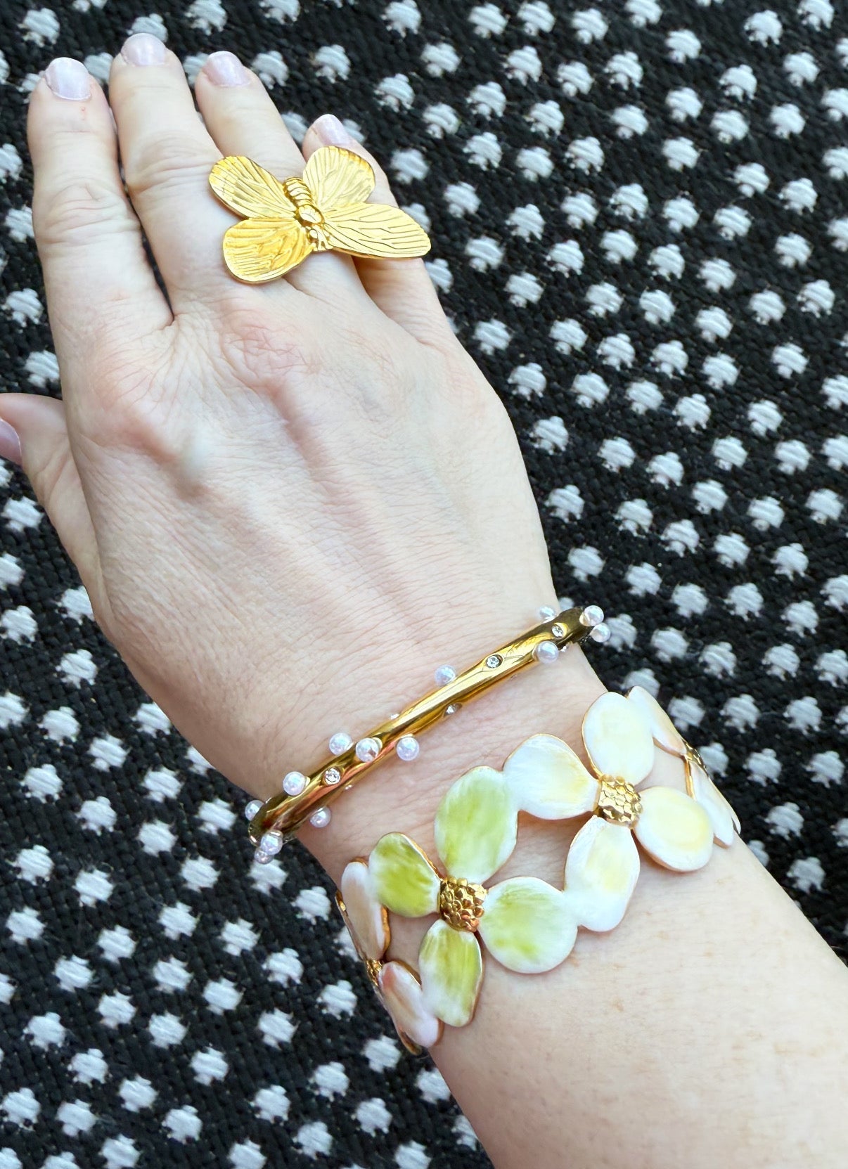 Hand wearing gold butterfly ring, a pearl and crystal gold bangle bracelet, and an enamel floral cuff on a black and white patterned background