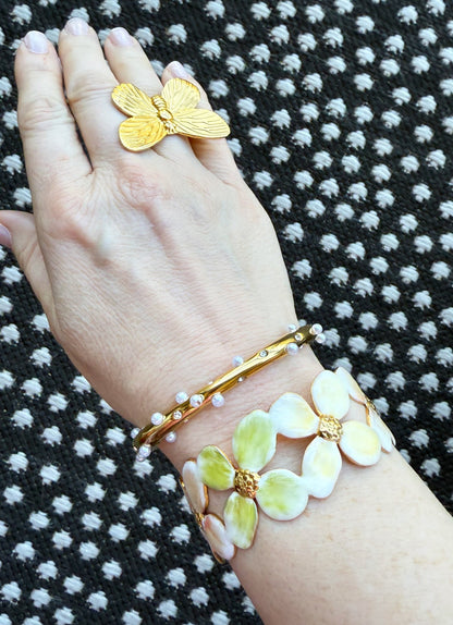 Hand wearing gold butterfly ring, a pearl and crystal gold bangle bracelet, and an enamel floral cuff on a black and white patterned background