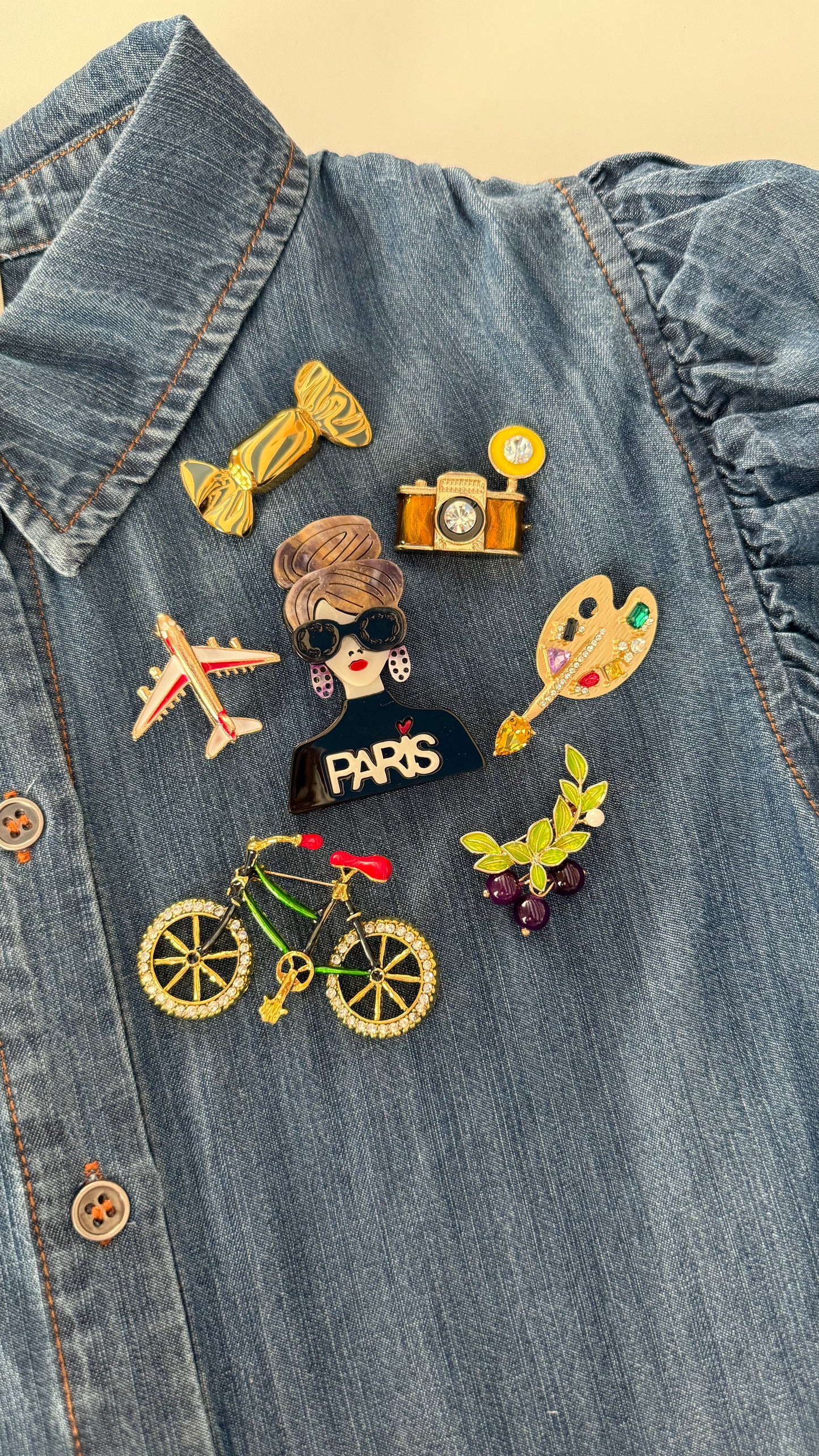 Collection of colorful pins on a denim jacket featuring Parisian brooches showing fashionista, camera, artist's palette, bicycle, airplane, candy, and grapes
