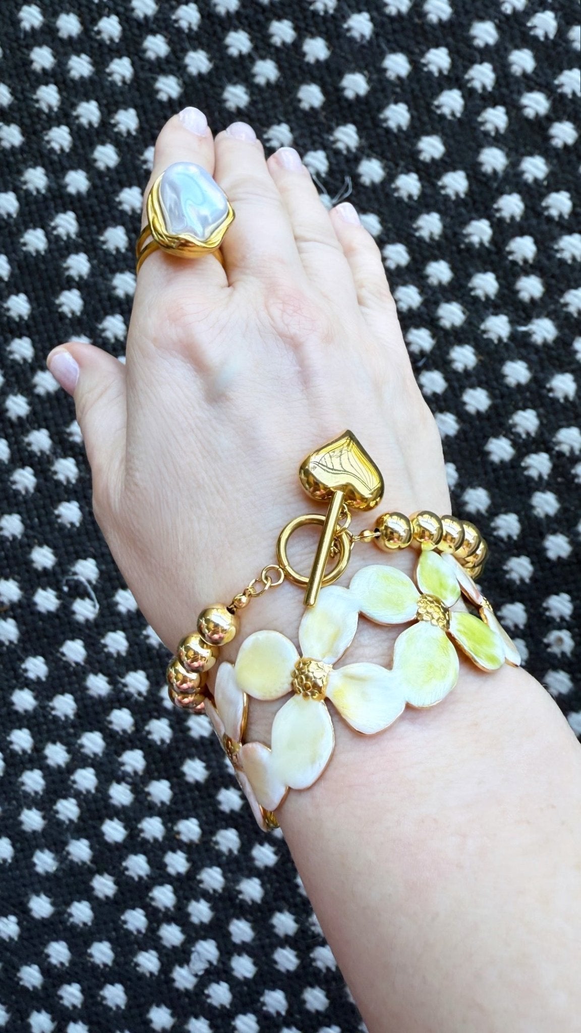 Hand wearing a pearl ring, gold bead heart charm bracelet, and a floral enamel cuff on a black and white patterned background