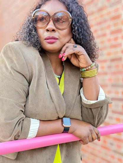 Woman wearing sunglasses and a beige jacket leaning on a pink hand railing outside with a brick wall background