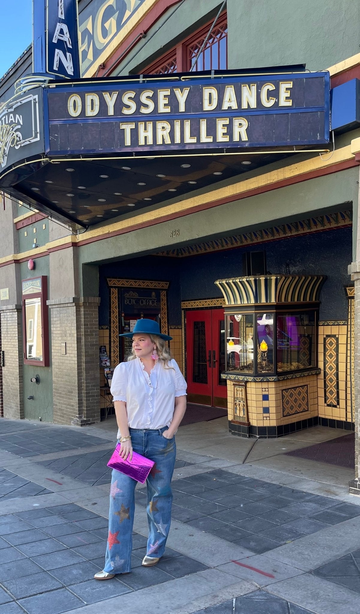 A woman standing in front of a theater with a marquee displaying 'Odyssey Dance Thriller' wearing star rhinestone embellished jeans
