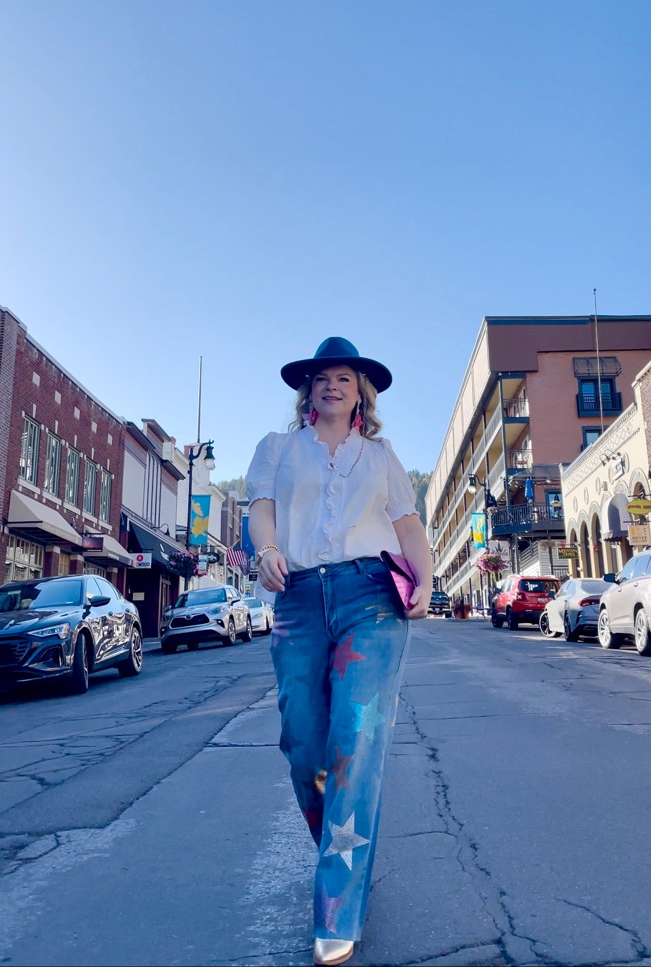 A woman walking on a city street wearing a white shirt, star rhinestone embellished jeans, and a hat.