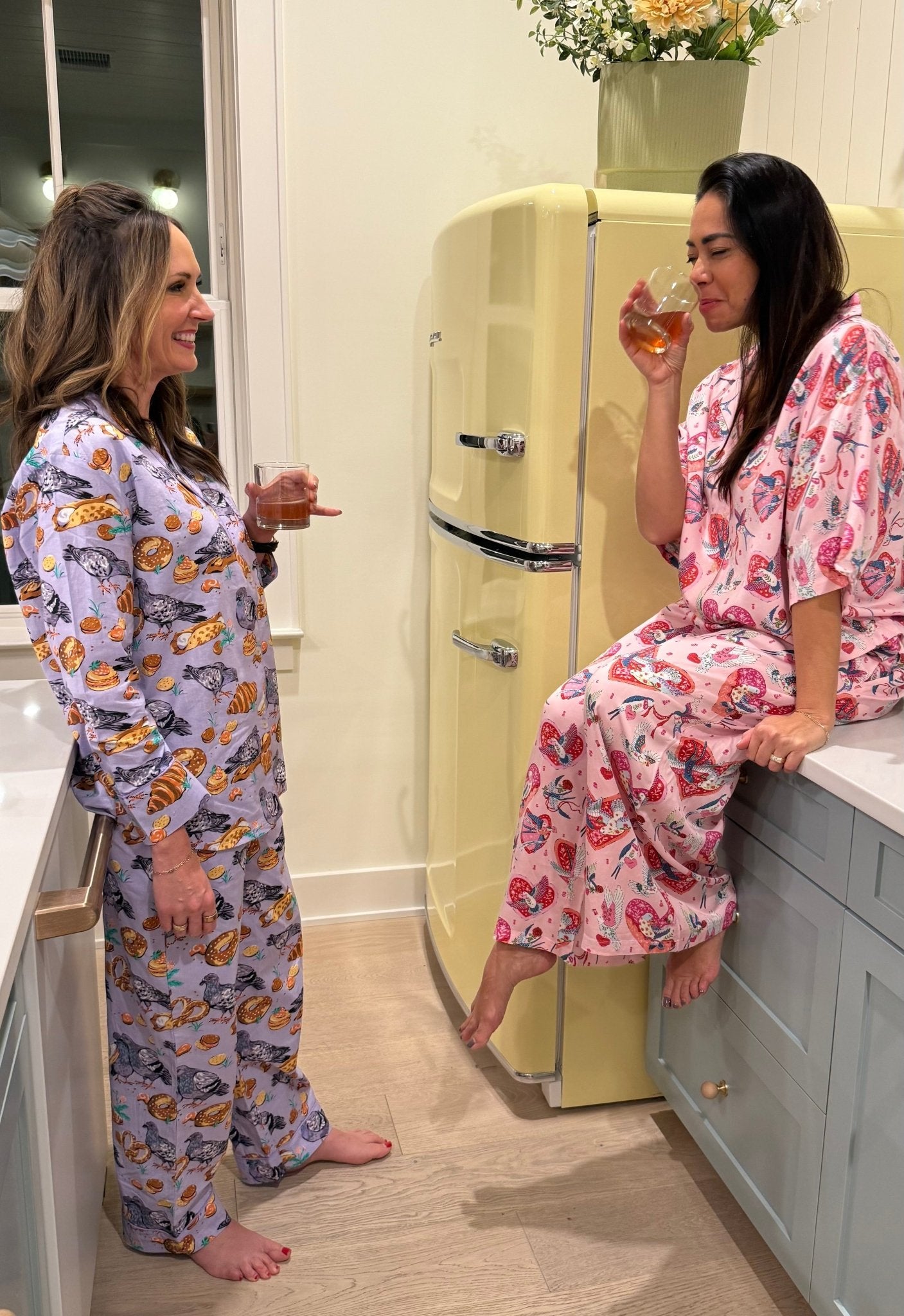 Two women in pajamas standing in a kitchen, one holding a glass of wine.