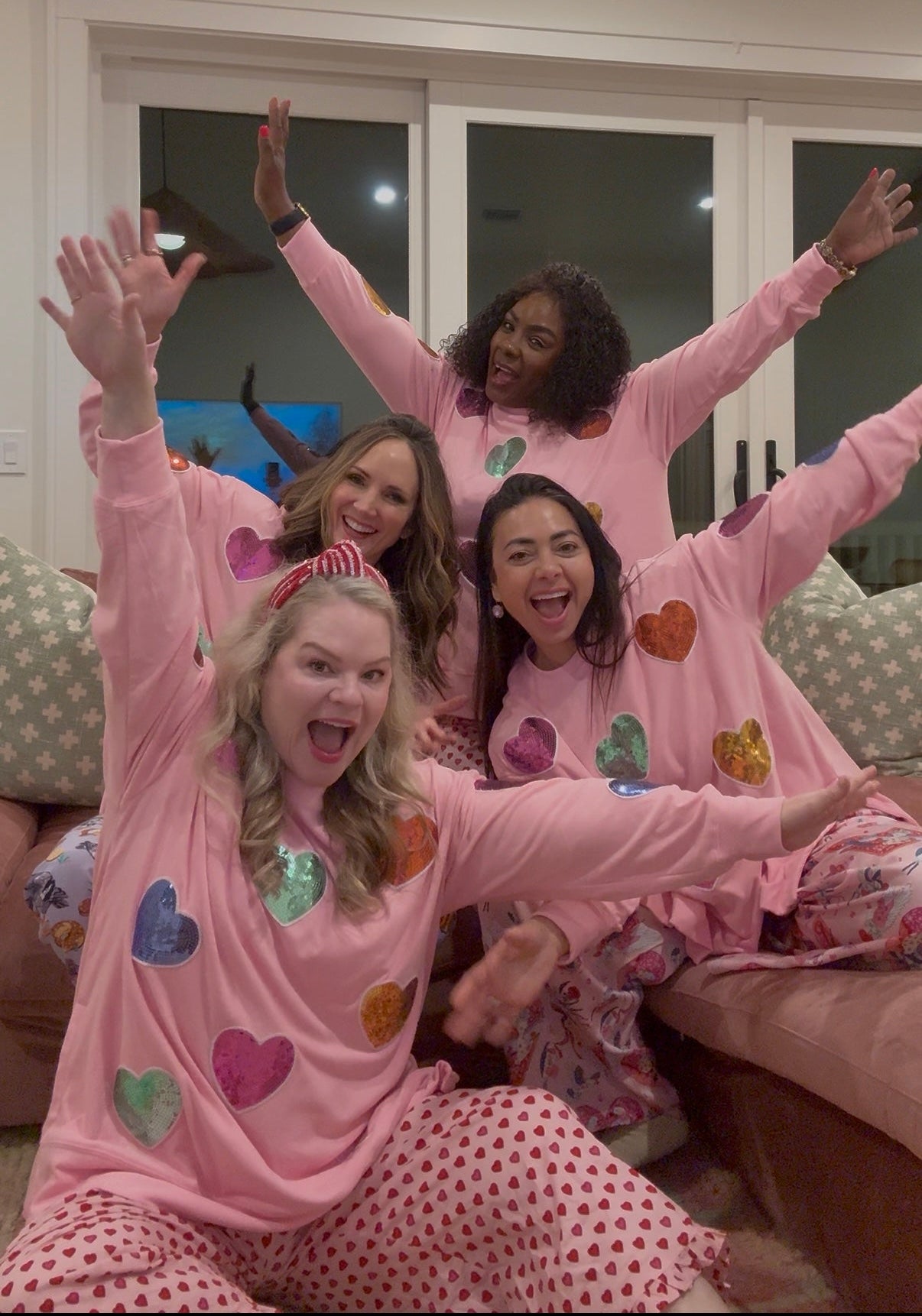 Four women in pink pajamas with heart patterns sitting on a couch with hands raised in the air.