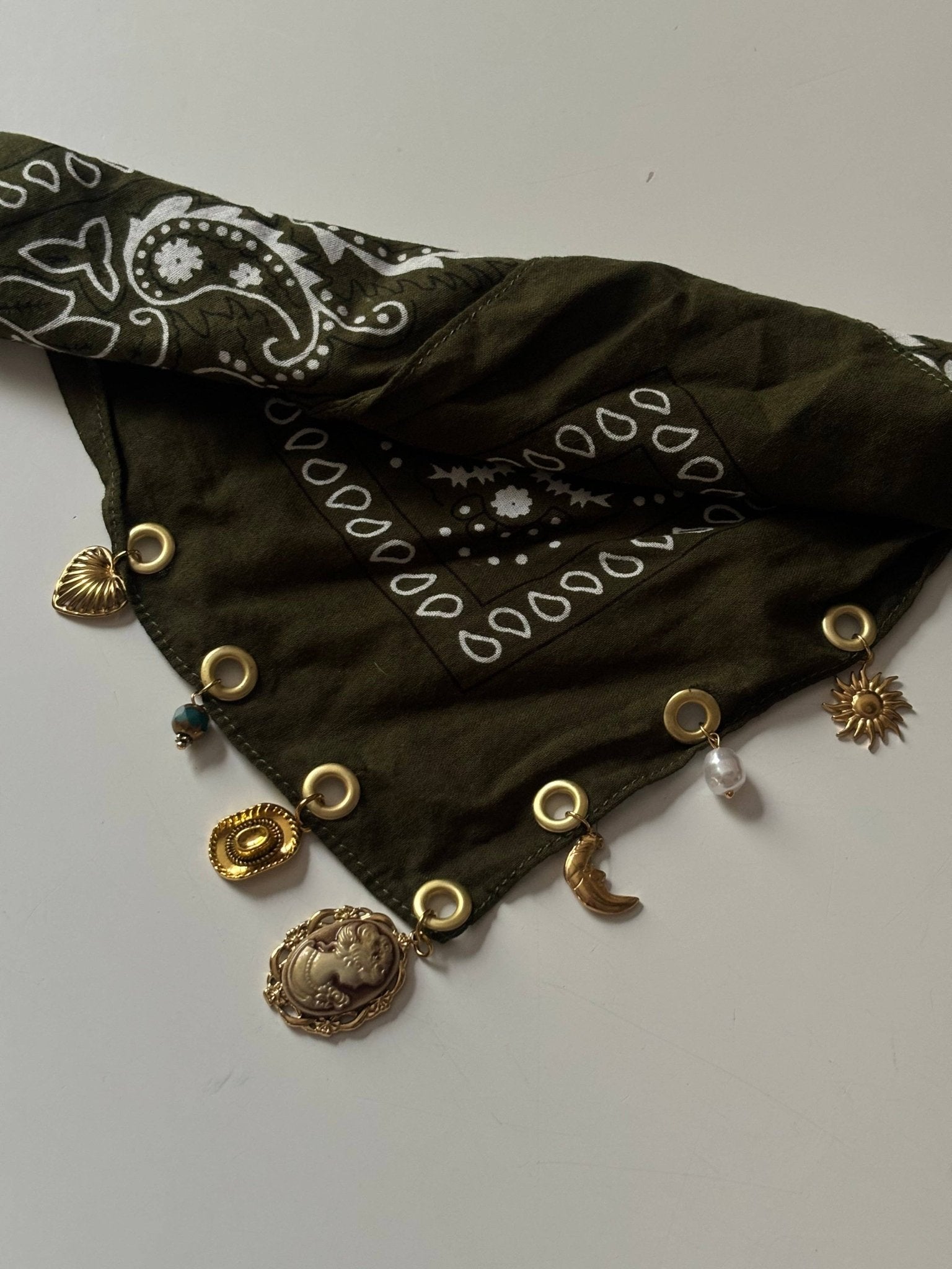 An olive green bandana with white paisley and star patterns, decorated with gold-tone metal grommets and various dangling charms including a crescent moon, sun, and medallion, laid flat on a light gray background.
