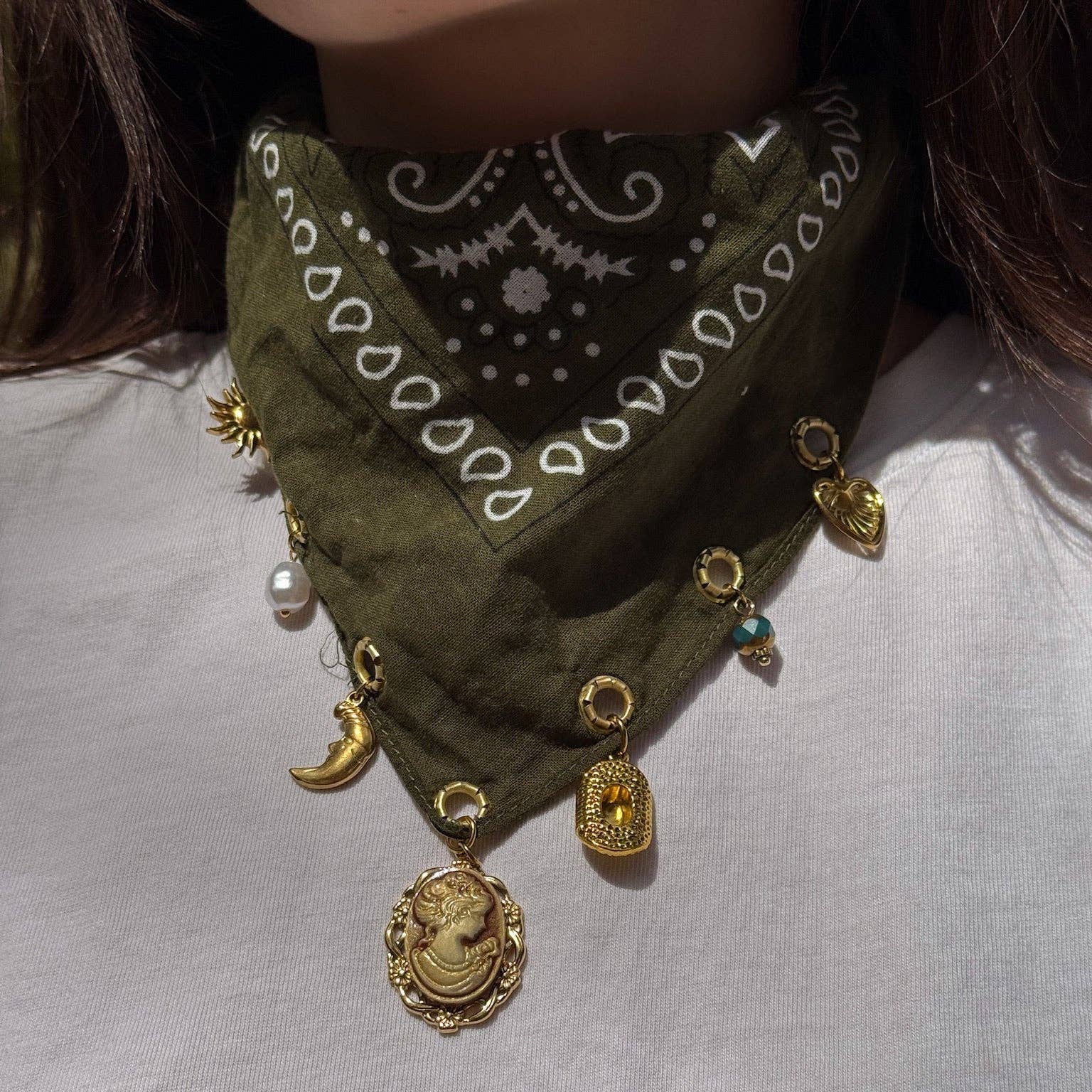 An olive green bandana bib necklace with white paisley patterns and multiple gold-toned charms, including a cameo bust, crescent moon, and medallion, worn by a person in a white shirt.