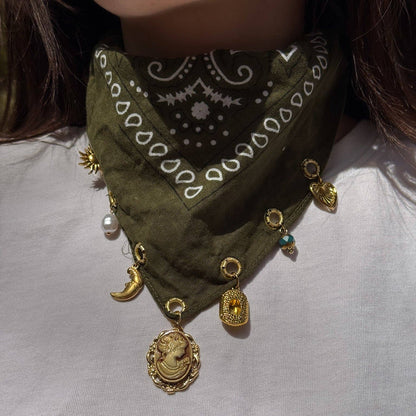 An olive green bandana bib necklace with white paisley patterns and multiple gold-toned charms, including a cameo bust, crescent moon, and medallion, worn by a person in a white shirt.