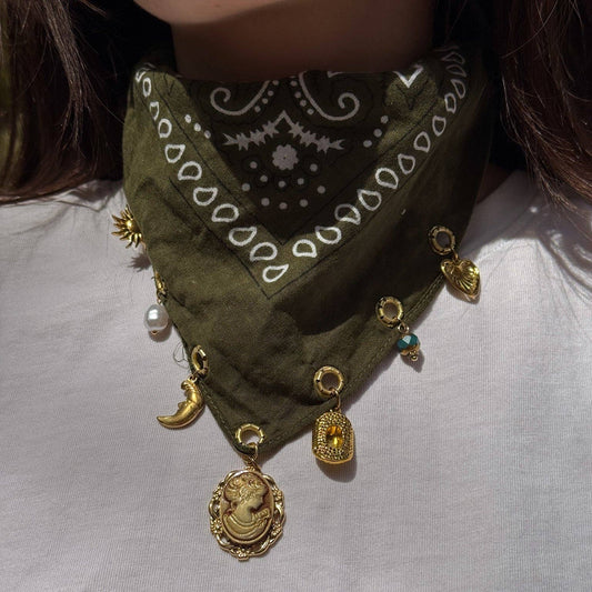 An olive green bandana bib necklace with white paisley patterns and multiple gold-toned charms, including a cameo bust, crescent moon, and medallion, worn by a person in a white shirt.