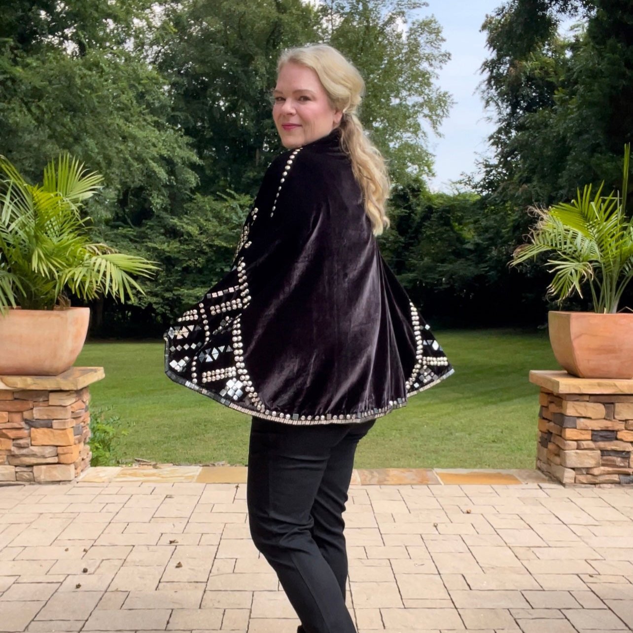 Woman wearing black velvet embellished capelet jacket with stud and mirror details showing back of capelet in an outdoor setting