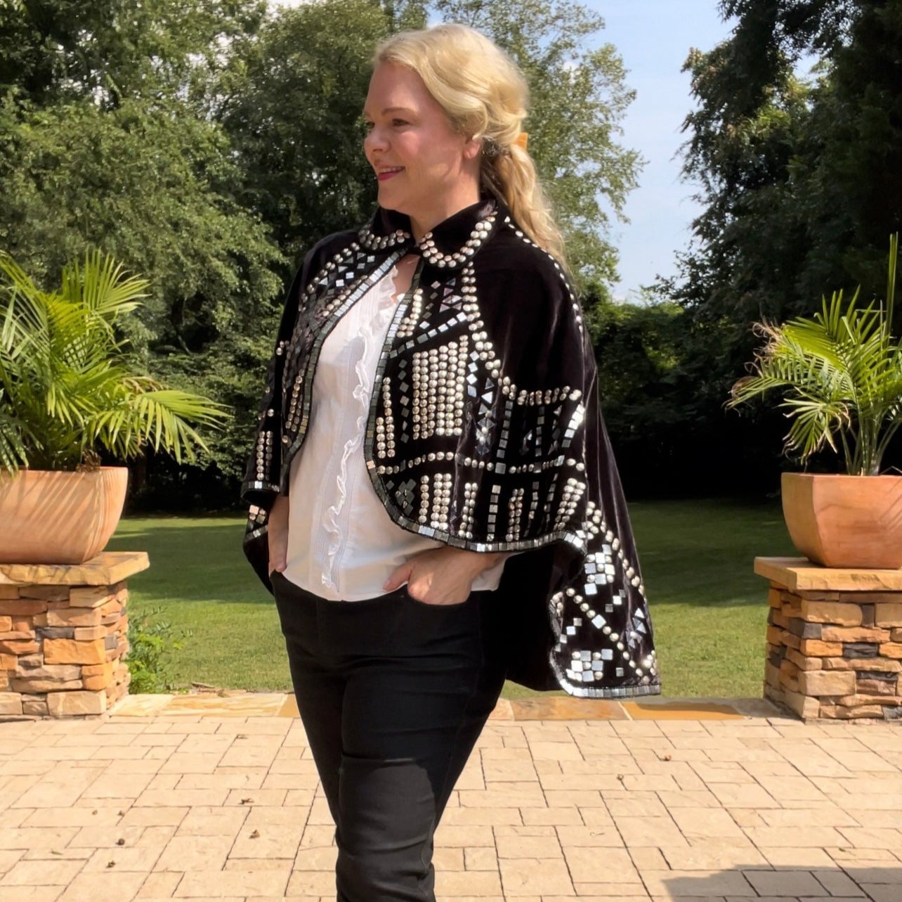Woman wearing black velvet embellished capelet jacket with stud and mirror details in an outdoor setting