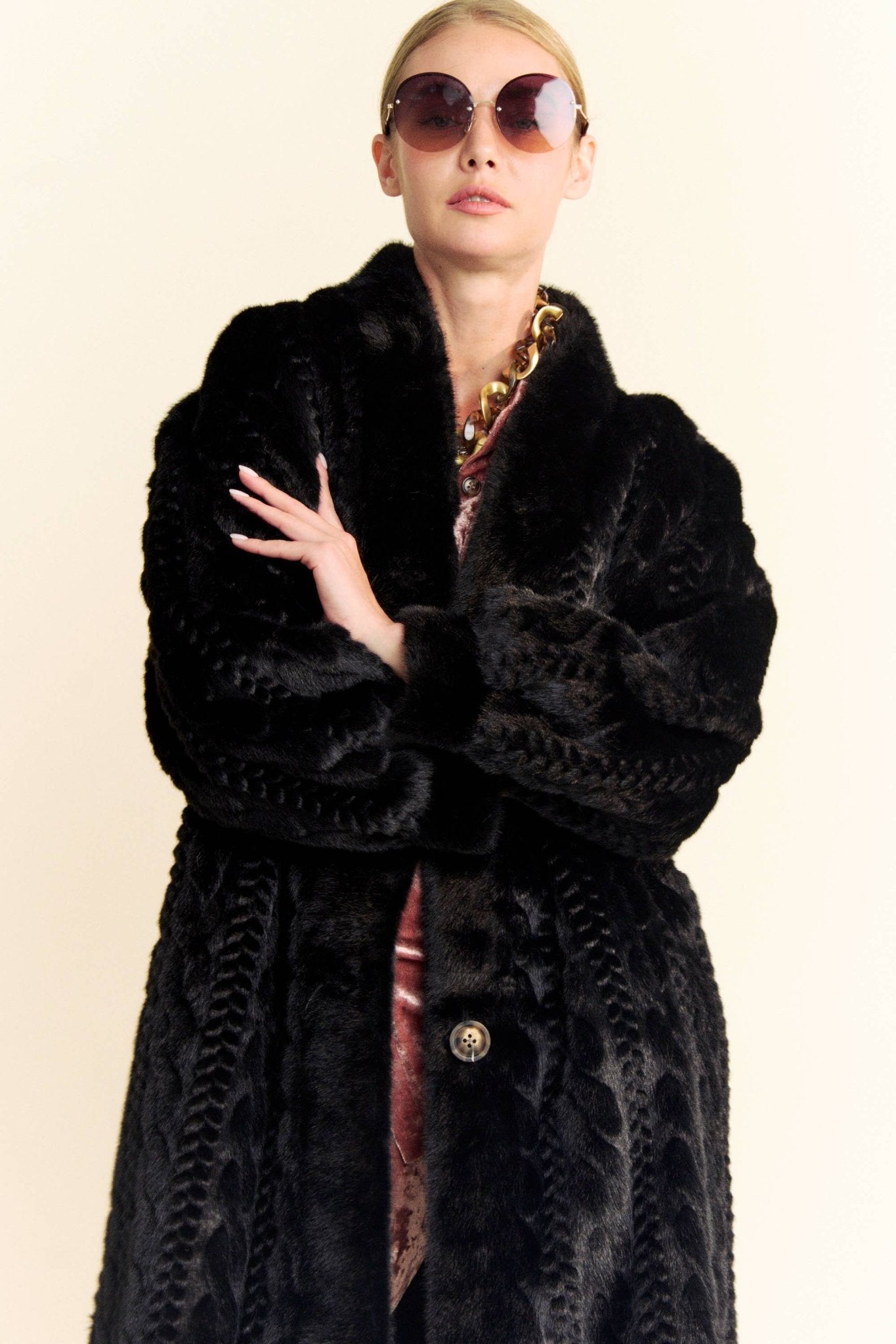 A woman in a black faux fur coat and round sunglasses against a white background