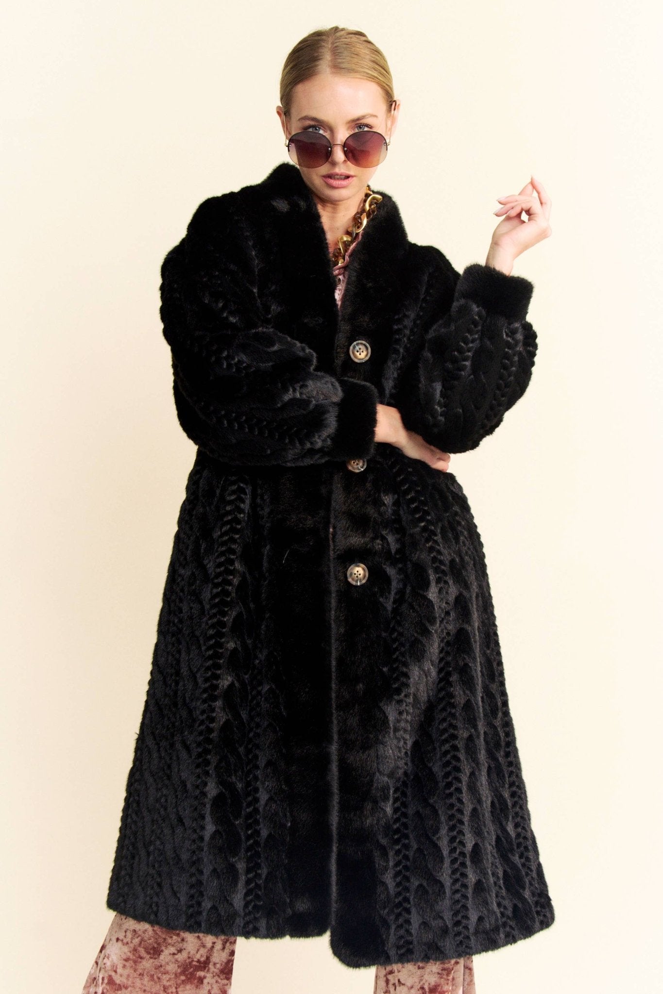 A model wearing a black faux fur coat with a stand-collar, featuring gold buttons. The model has sunglasses and is posing with one hand on her hip.
