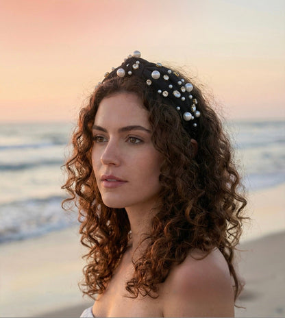 Woman with curly hair wearing a black headband with pearls against a beach sunset.