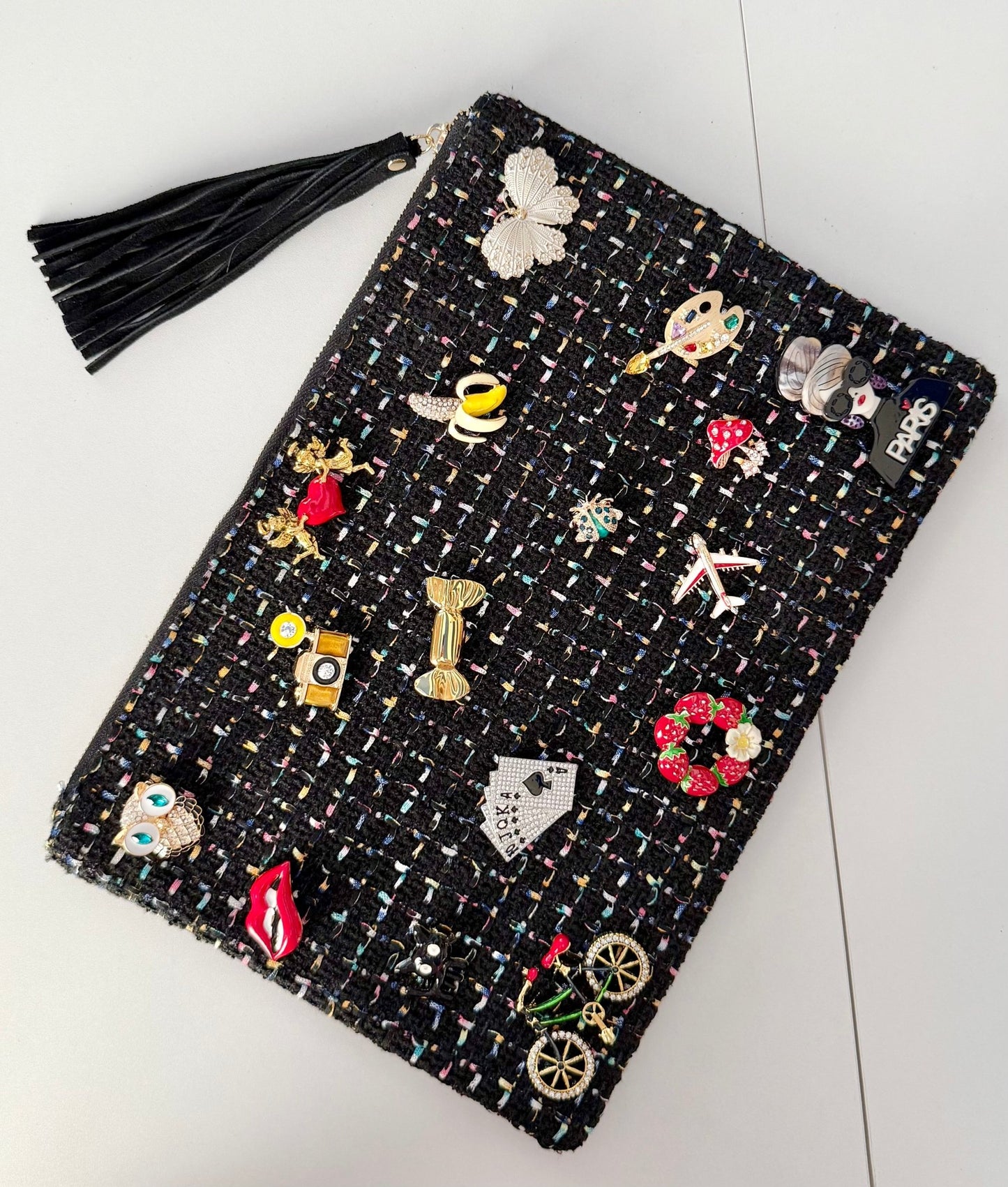 Black tweed pouch with 18 colorful brooches including owl, lips, and Paris designs — front view on gray background