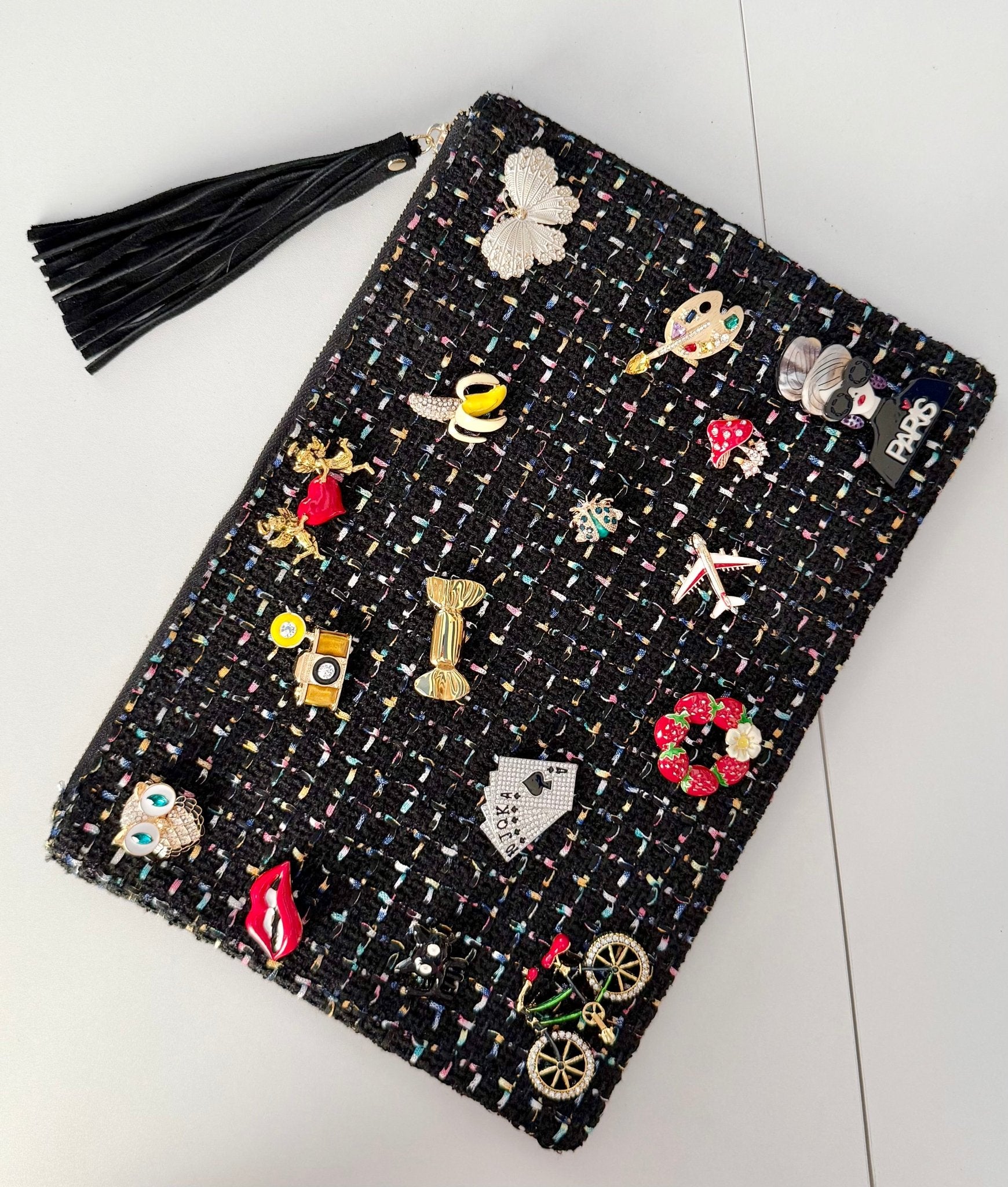 Black tweed pouch with 18 colorful brooches including owl, lips, and Paris designs — front view on gray background