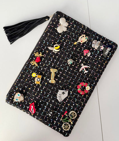 Black tweed pouch with 18 colorful brooches including owl, lips, and Paris designs — front view on gray background