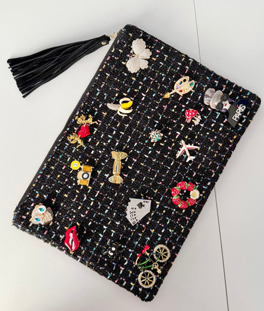 Black tweed pouch with 18 colorful brooches including owl, lips, and Paris designs — front view on gray background