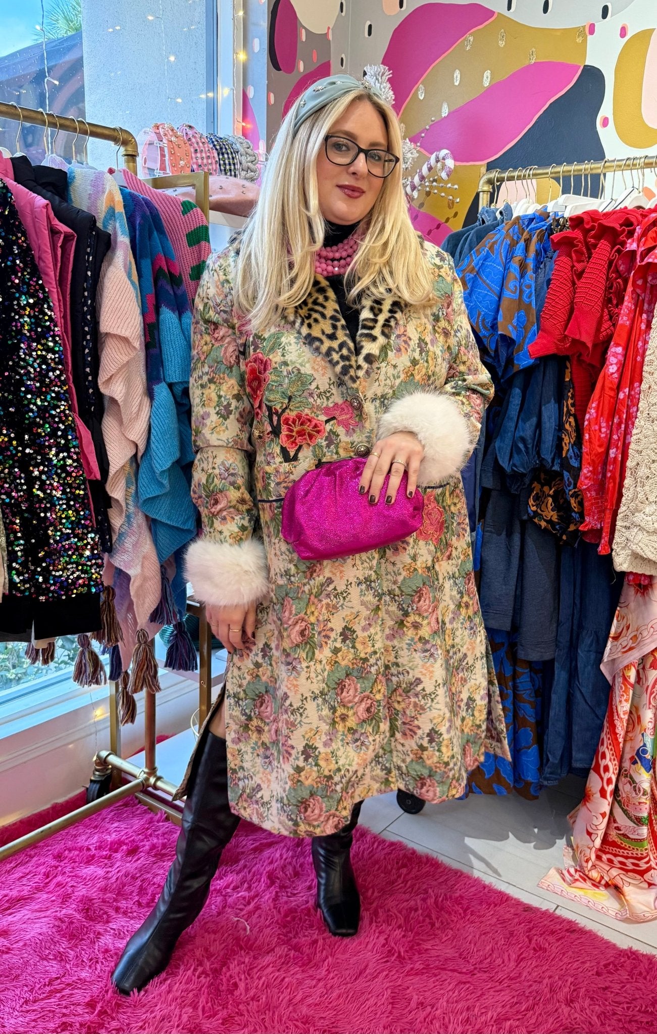 Woman in a floral coat with fur trim, light blue crystal embellished headband, and pink clutch standing in front of a clothing rack inside a boutique