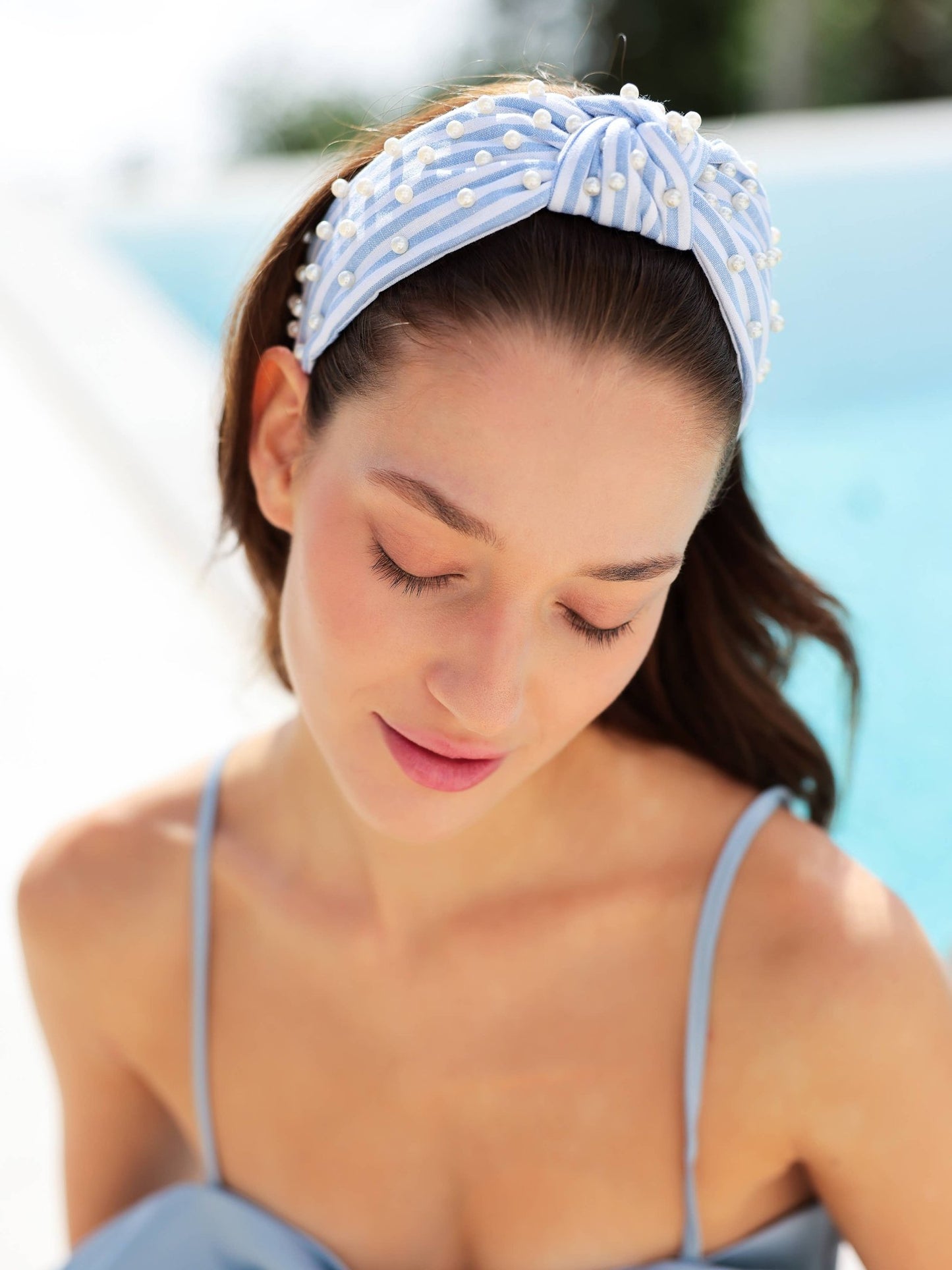 Woman wearing a light blue headband with white polka dots and pearls, standing against a blurred outdoor background.
