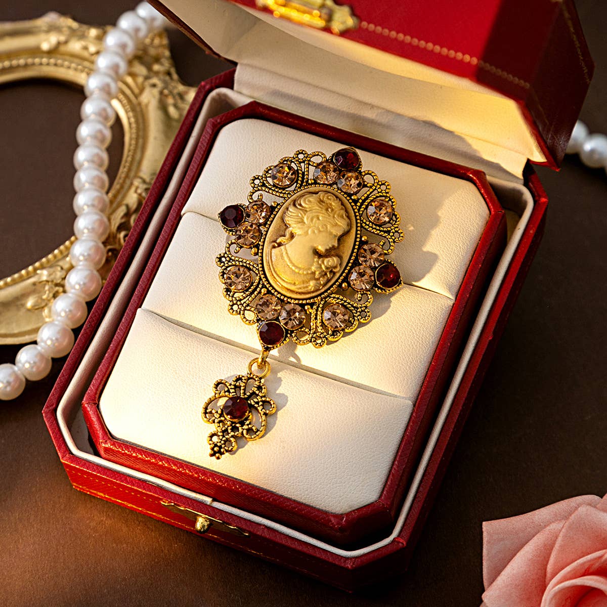 Gold brooch with gemstones and a center cameo in a red jewelry box on a dark surface.