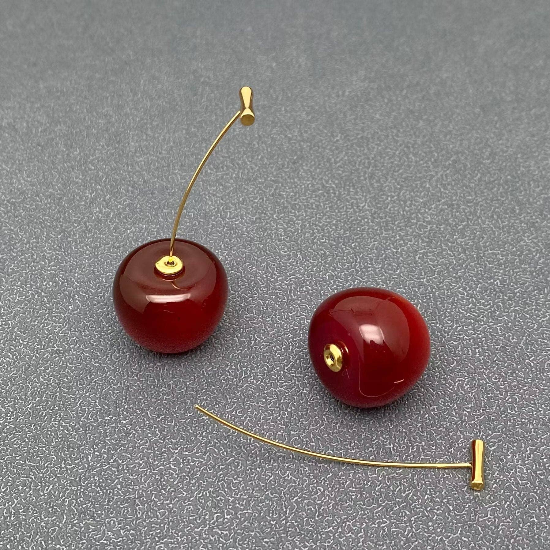 a pair of red cherry resin earrings with gold stems laying on a grey background