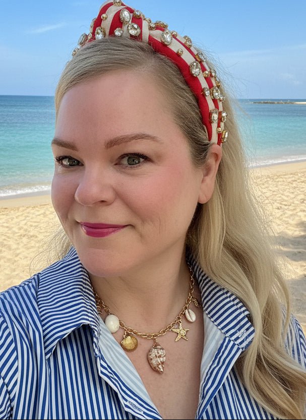 Woman wearing a red headband with gold accents and a necklace with various ocean themed charms with a beach background
