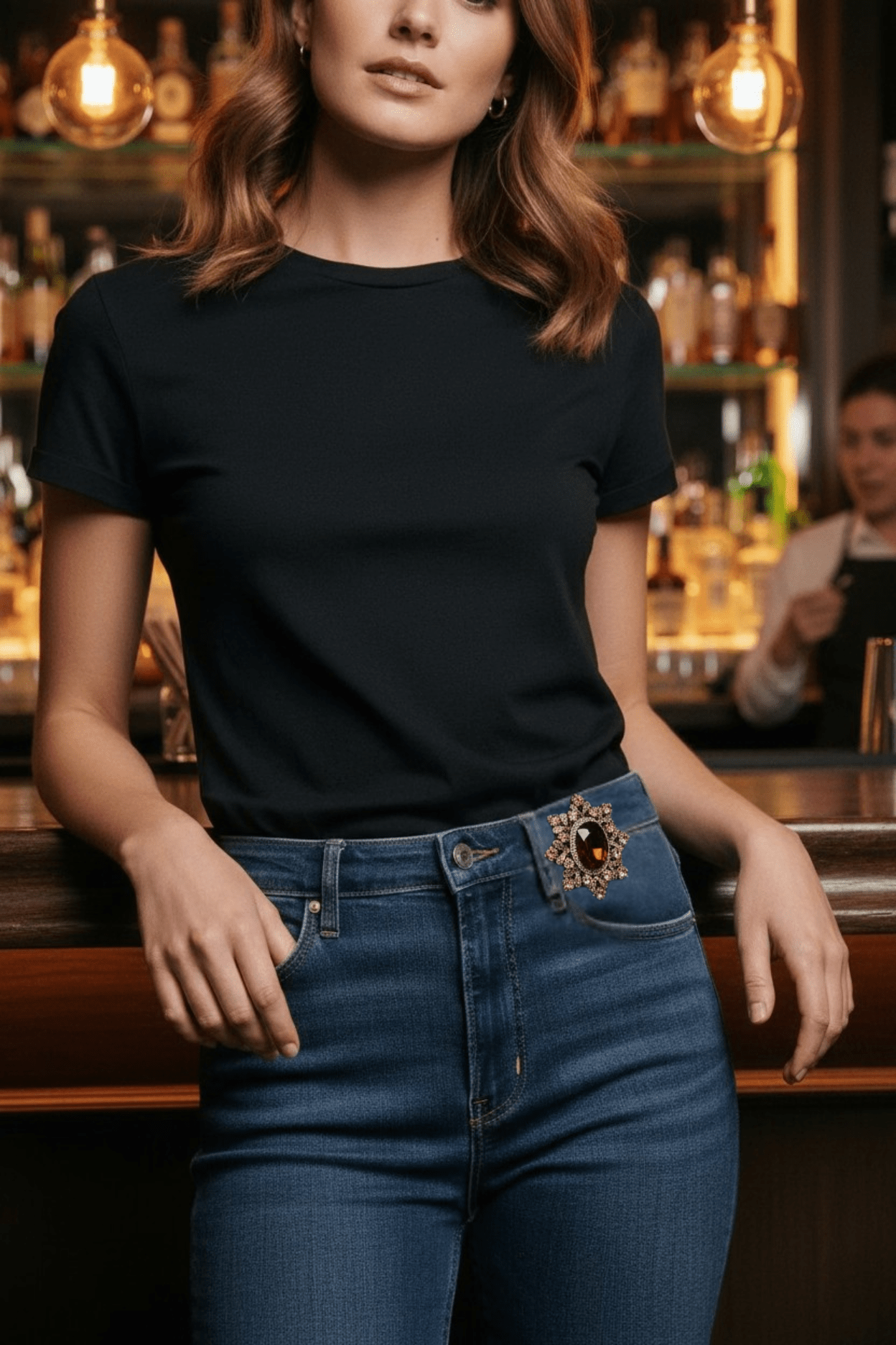 Woman wearing a black t-shirt and blue jeans with a decorative brooch in a bar setting.