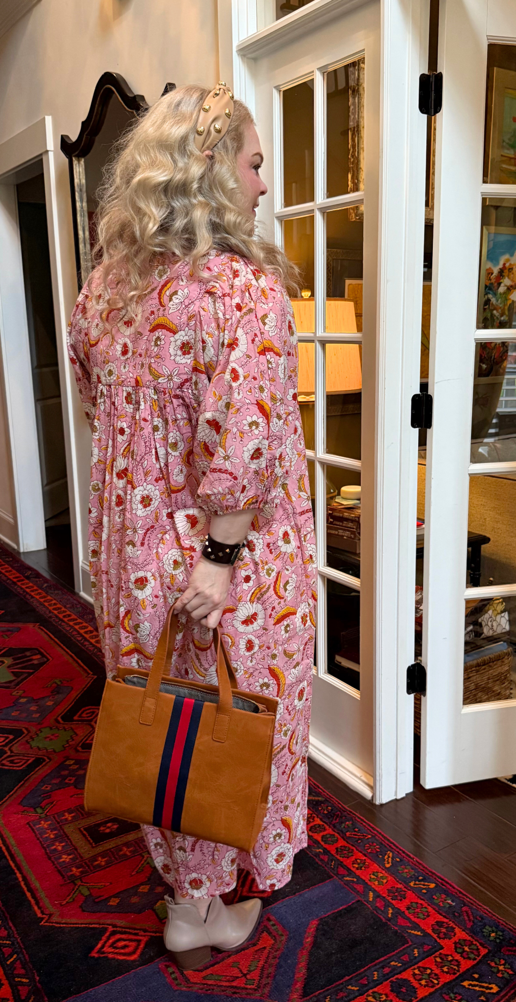Back view of a woman in a pink floral dress holding a brown handbag with a red and navy stripe, standing in a home interior.