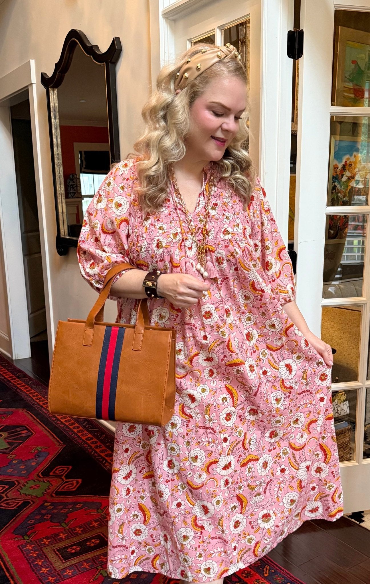 Woman in a pink floral dress holding a brown handbag with a red and navy stripe, standing in a home interior.