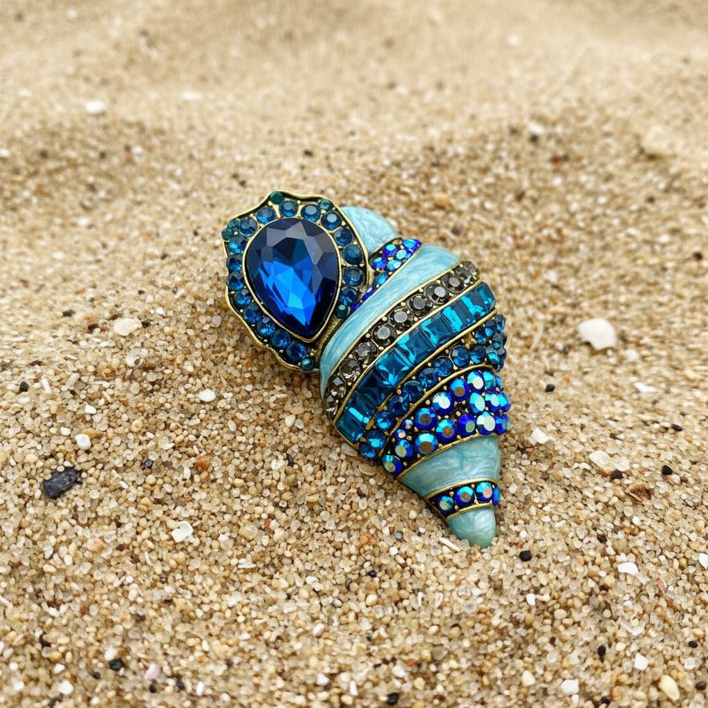 Blue enamel conch shell brooch with rhinestone crystal detail on sand — ocean-inspired statement jewelry pin