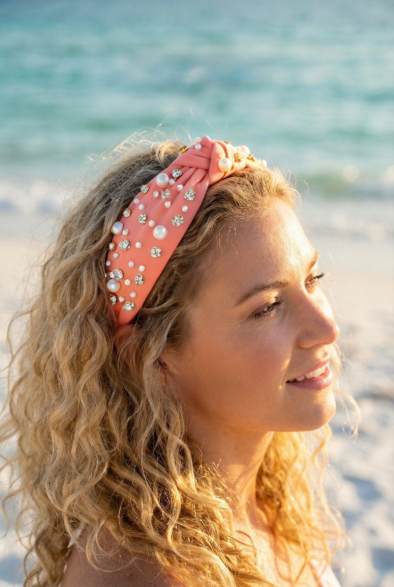 Woman with blonde curly hair wearing a decorative coral colored headband on a beach.