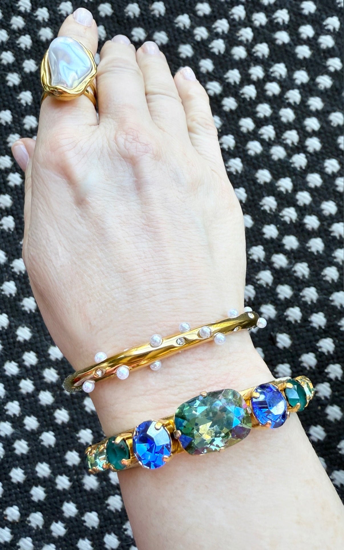 Hand wearing a gold pearl ring, a pearl and crystal embellished bracelet, and a bracelet with gemstones on a textured black and white background