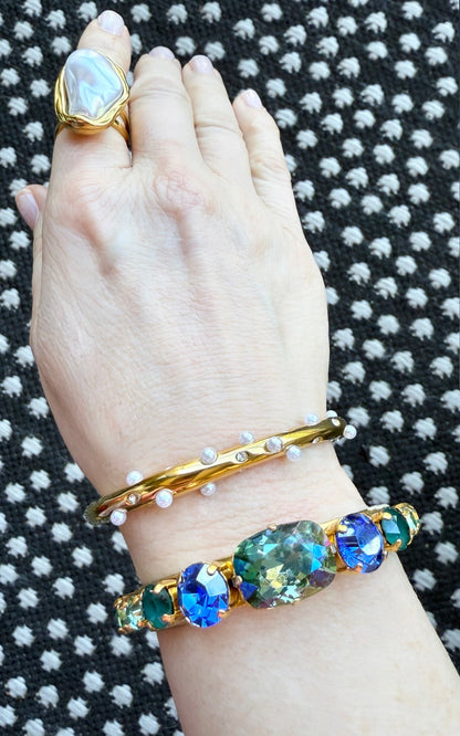 Hand wearing a gold pearl ring, a pearl and crystal embellished bracelet, and a bracelet with gemstones on a textured black and white background