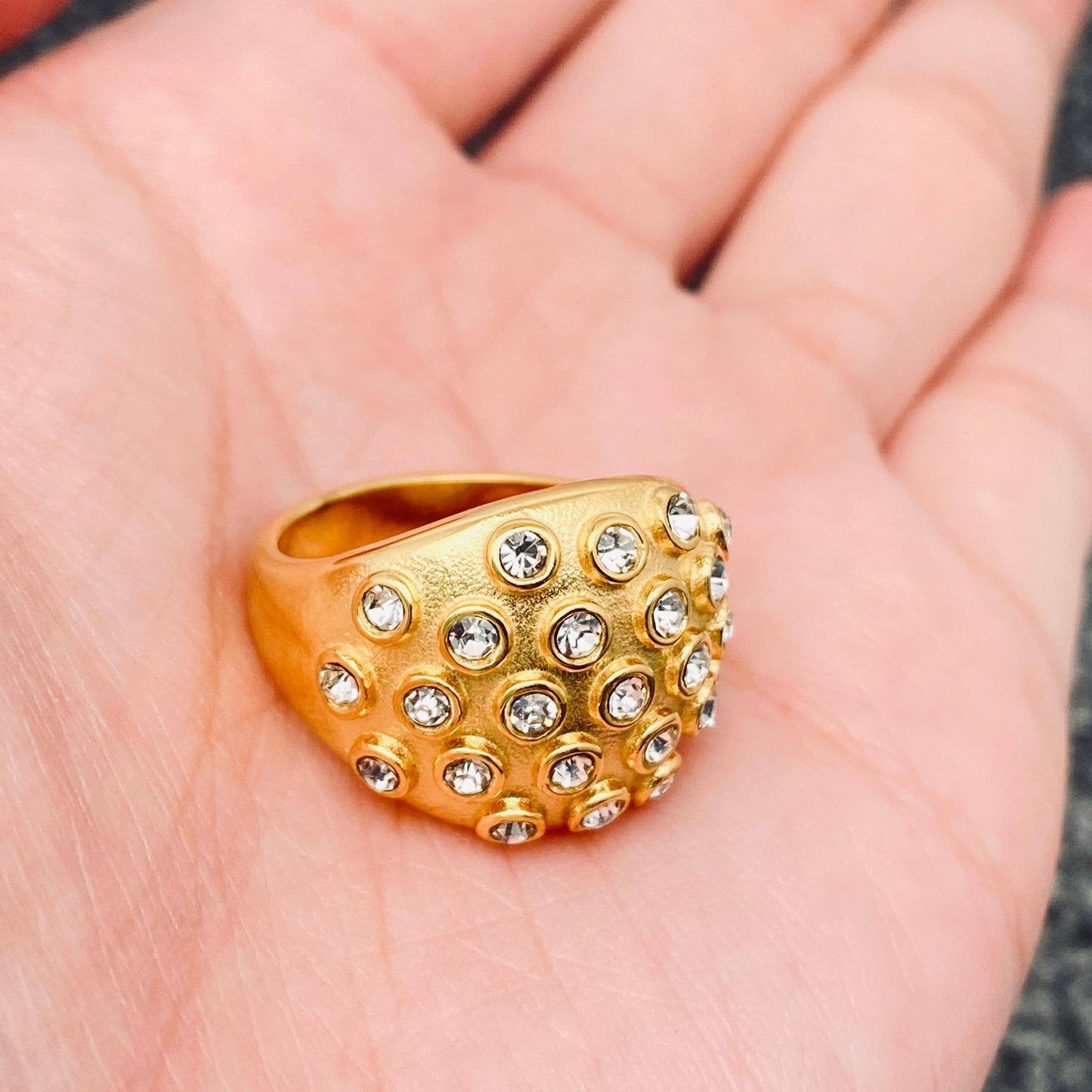 A hand holding a gold statement ring with clear crystal stones on top.