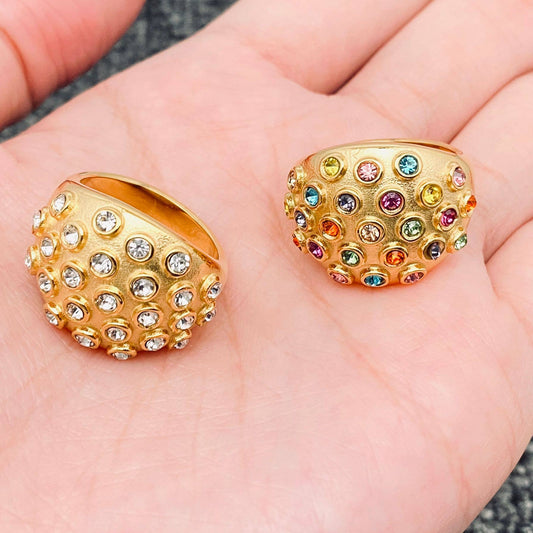 A hand holding 2 crystal dome statement rings in gold. One ring has clear crystal stones and the other ring and multi colored stones.