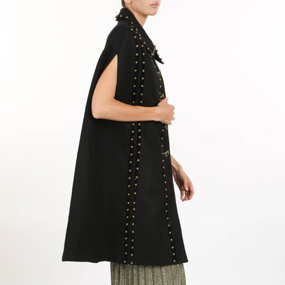 side view of a woman wearing a black cape coat with gold stud detailing against a white background