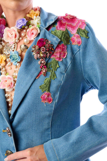a close up view of a blue denim jacket with floral embroidery and a crystal brooch on the lapel against a white background
