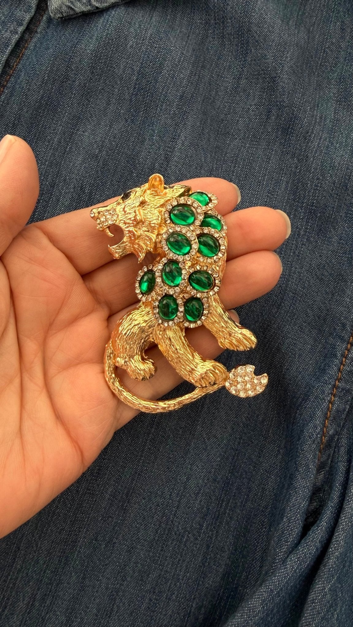 Gold and green jeweled lion brooch held in a hand against a denim background