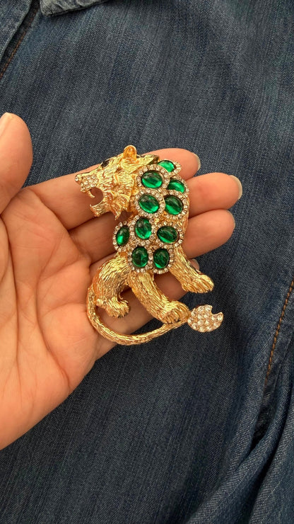Gold and green jeweled lion brooch held in a hand against a denim background
