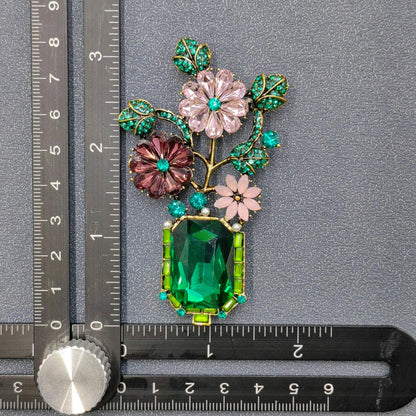 Decorative brooch with flowers and green gemstone next to a ruler for scale on a gray background