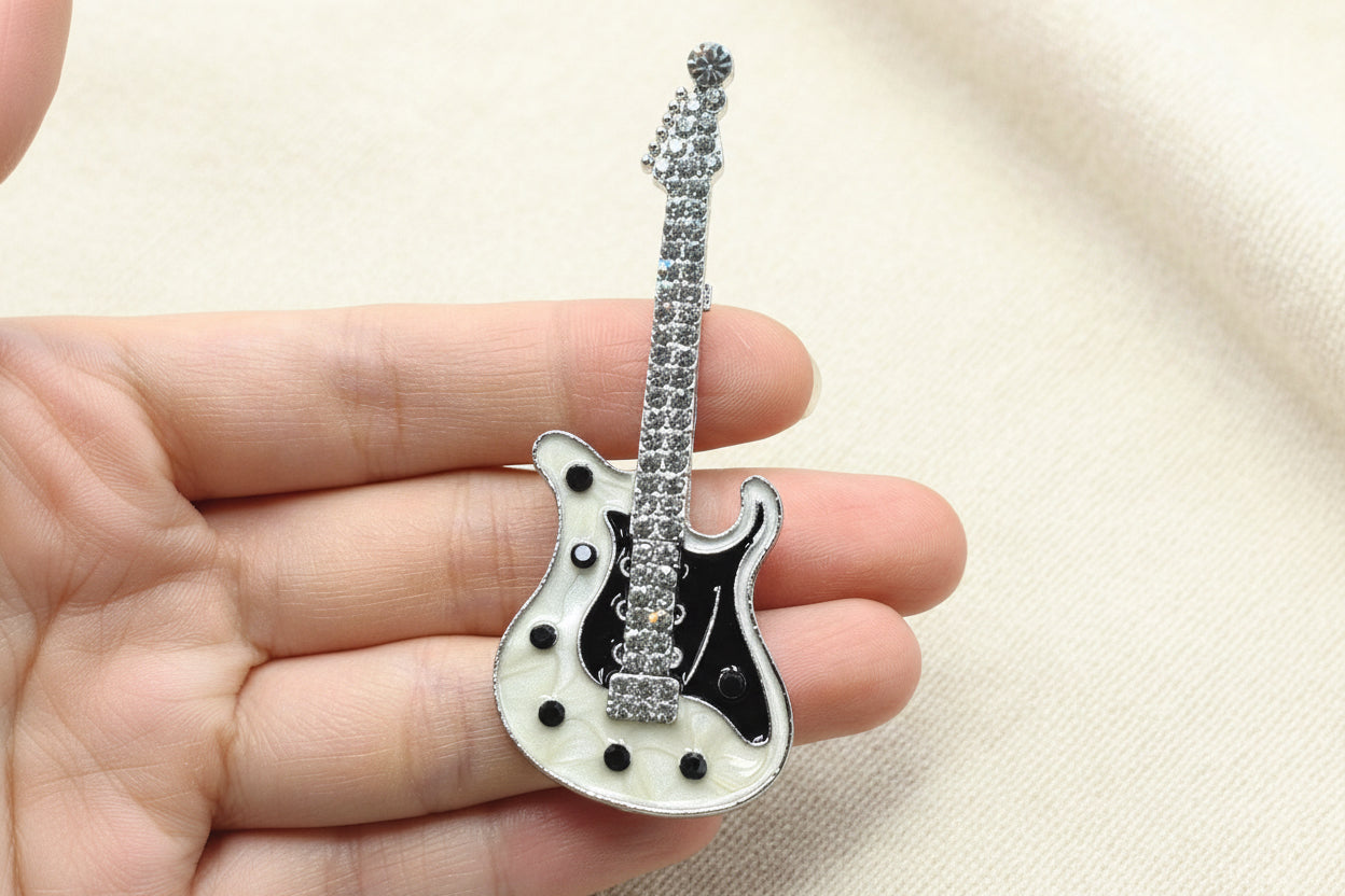 Guitar brooch with white and black enamel body and clear crystals held in a hand against a grey background