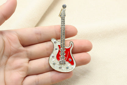 Decorative guitar-shaped brooch with red and white body and clear crystal details held in a hand against a gray background