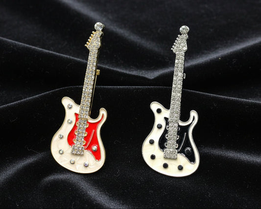 Two guitar brooches- one is white & red enamel with clear crystals and the other one is white & black enamel with clear crystals against a black velvet fabric background