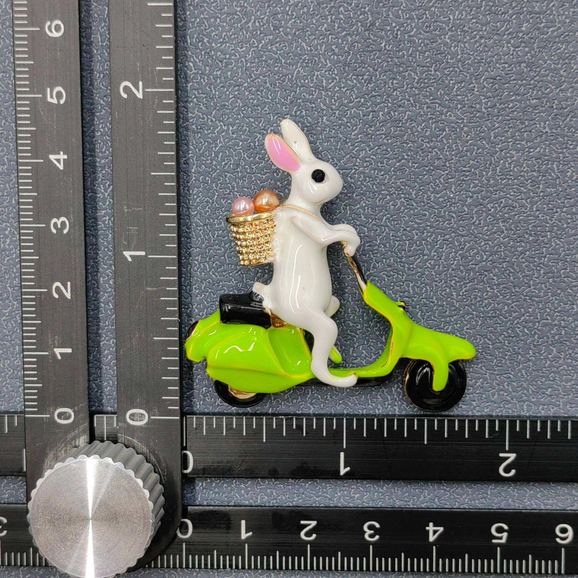 White rabbit on a green scooter brooch with a basket of eggs, next to a ruler for scale.