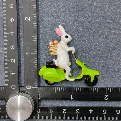 White rabbit on a green scooter brooch with a basket of eggs, next to a ruler for scale.
