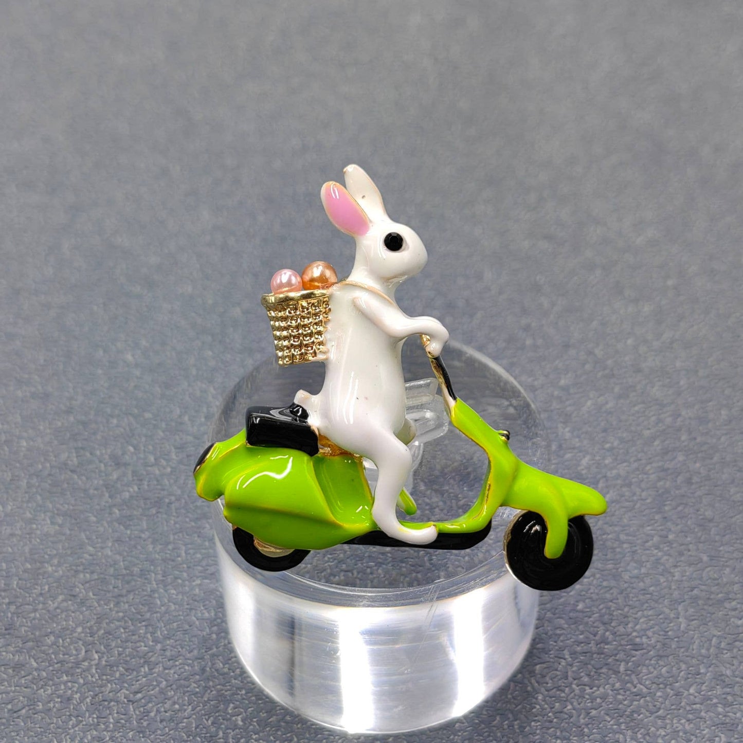White rabbit brooch on a green scooter with a basket of eggs on a gray background