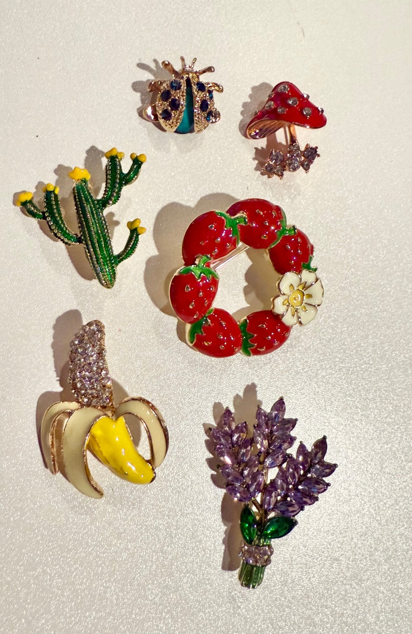 Set of colorful brooches including a cactus, strawberry wreath, banana, and flower arrangement on a light background.